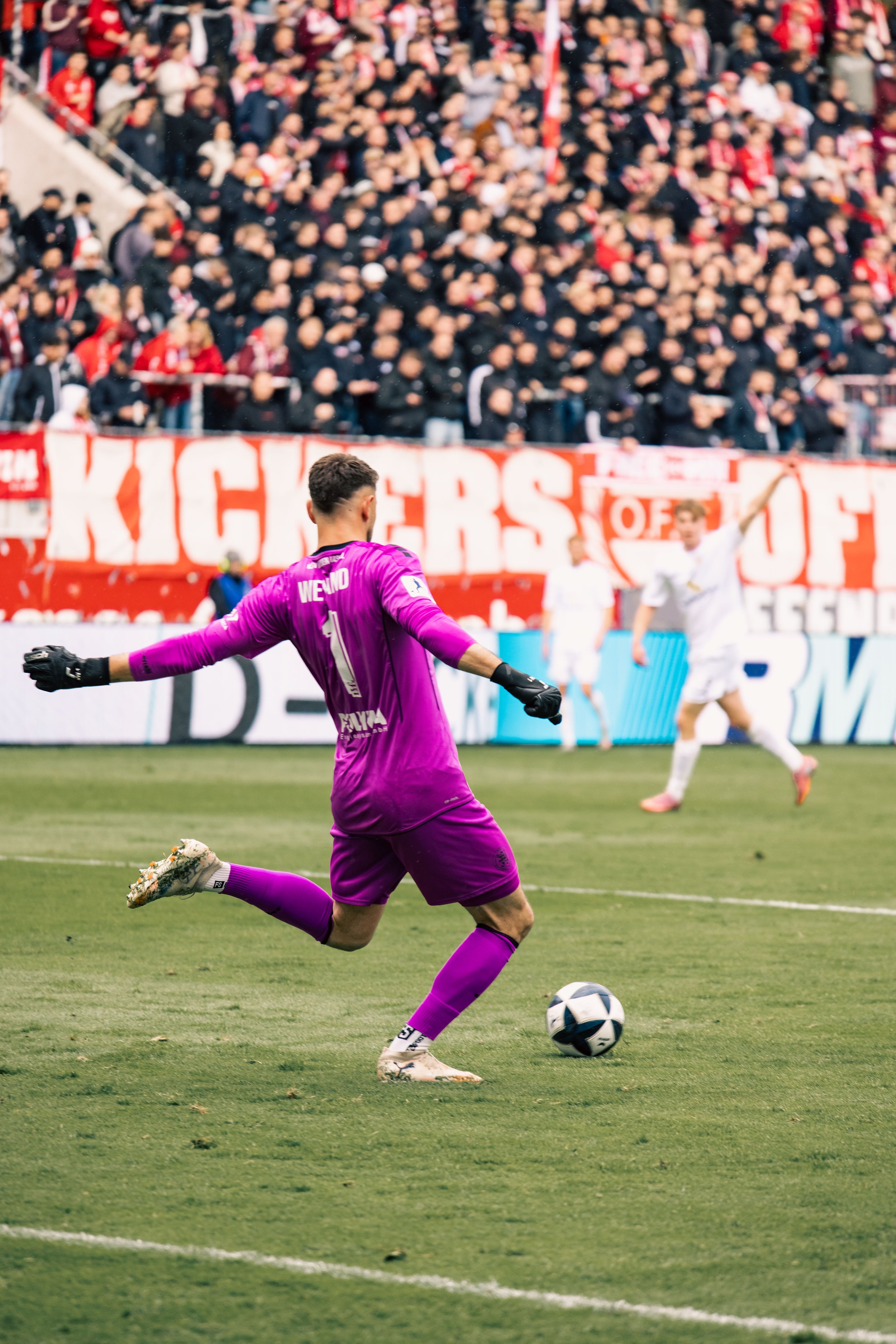 Fußballspieler in lila Trikot schießt auf den Ball während ein Fanblock mit roten und weißen Bannern im Hintergrund zu sehen ist.