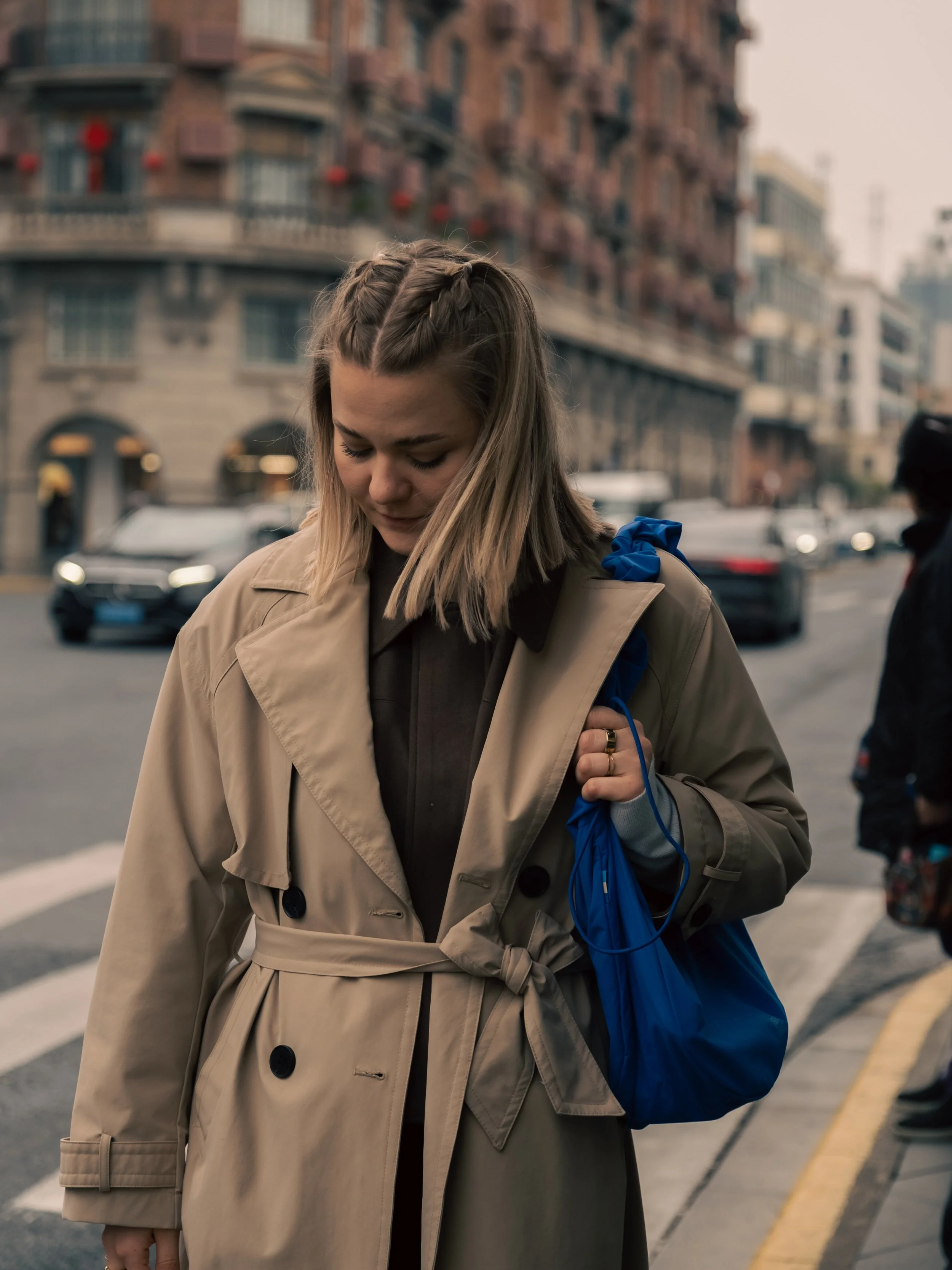 DominoKati mit hellbraunen Haaren und Zopf trägt einen beige Trenchcoat in Shanghai und hält eine blaue Tasche auf einer Stadtstraße. Im Hintergrund sind mehrstöckige Gebäude und Autos sichtbar.