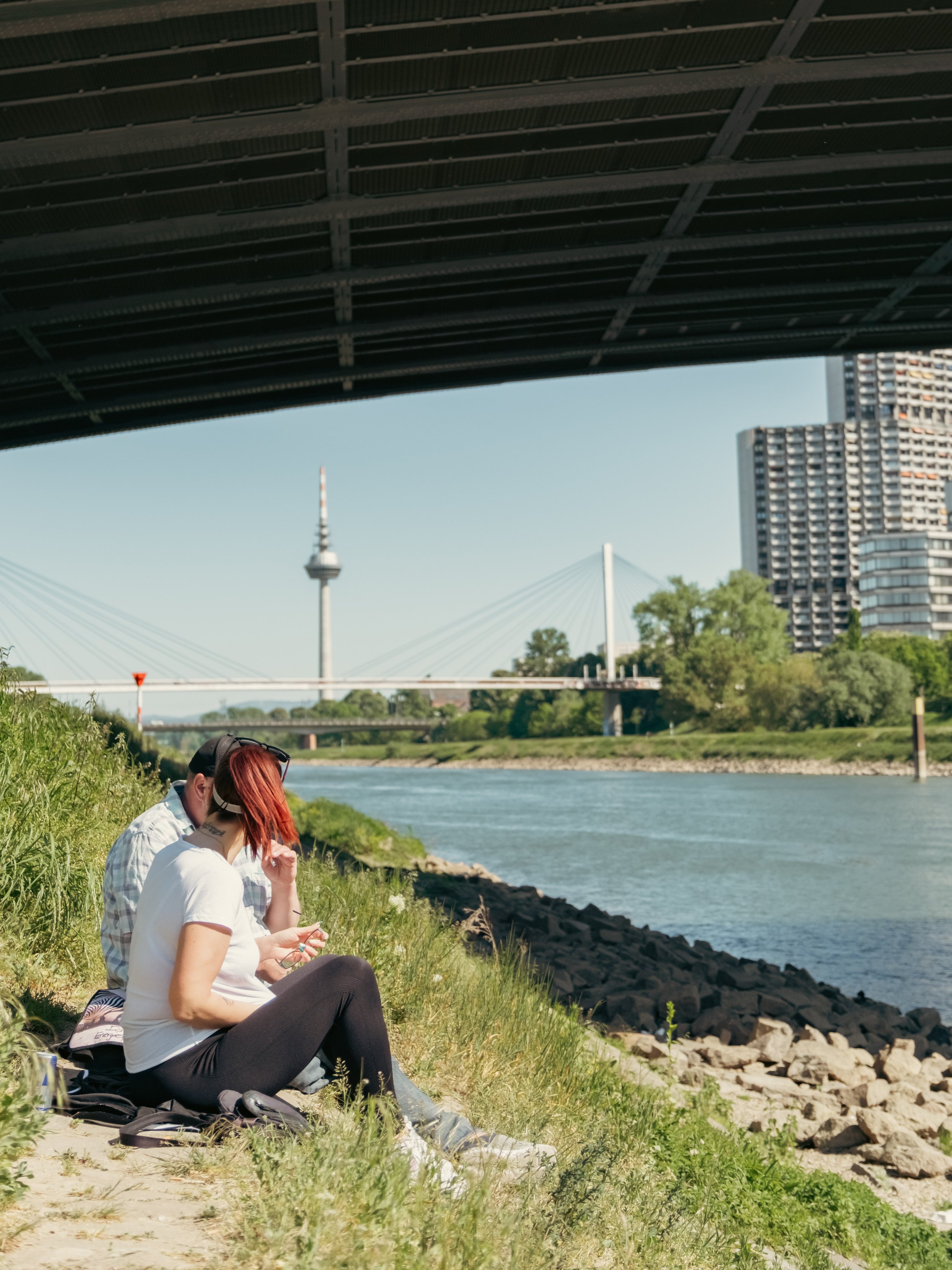 Zwei Menschen sitzen am Ufer eines Flusses, unter einer Brücke, im Hintergrund der Fernmeldeturm von Mannheim, eine Brücke und Hochhäuser sichtbar.
