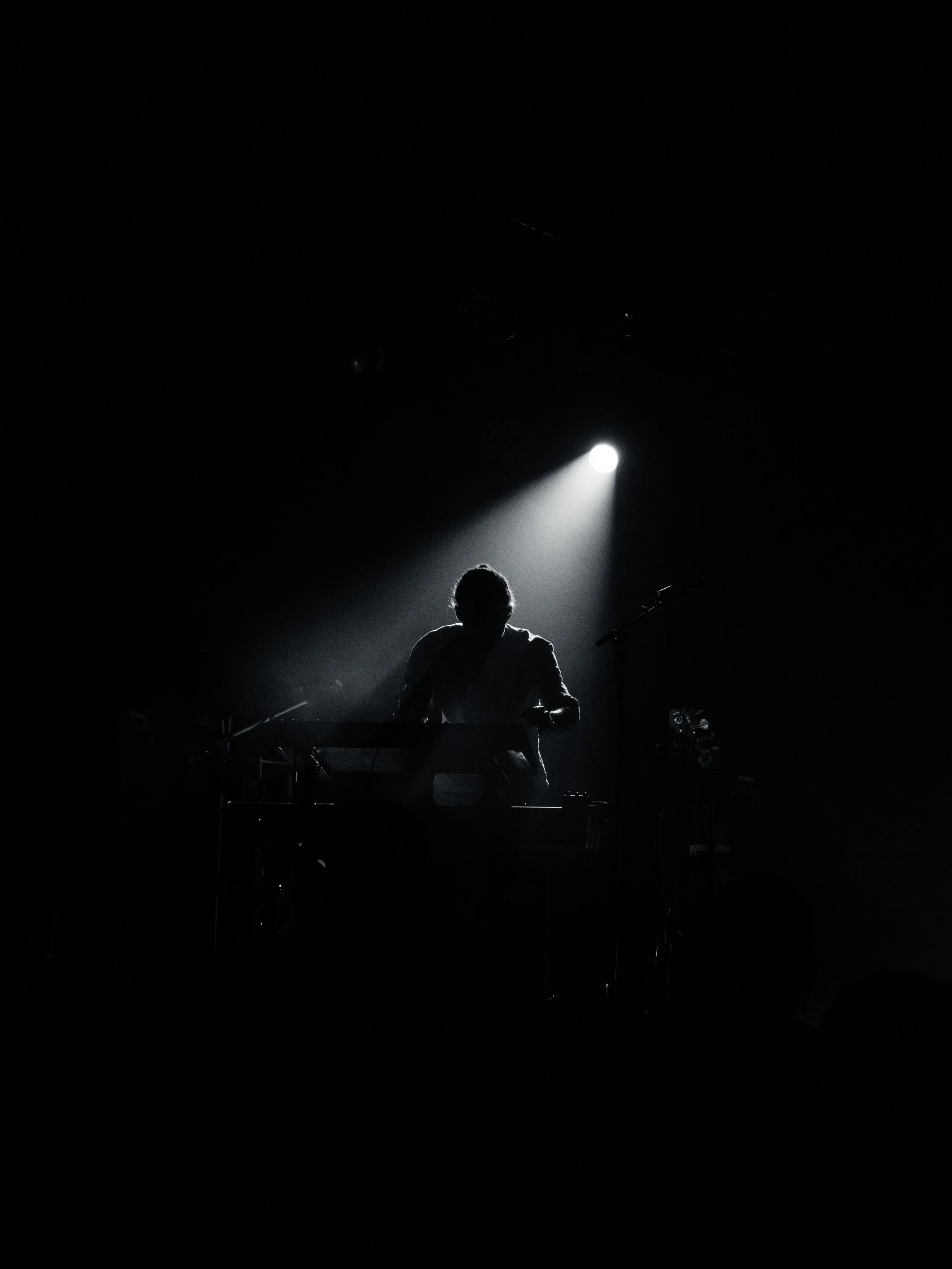 Eine Person sitzt auf einem Tisch mit einem Keyboard, beleuchtet von einem fokussierten Lichtstrahl in einer dunklen Umgebung. Event-Fotografie bei einem Konzert im Zoom, Frankfurt. Fotografiert aus dem Publikum.