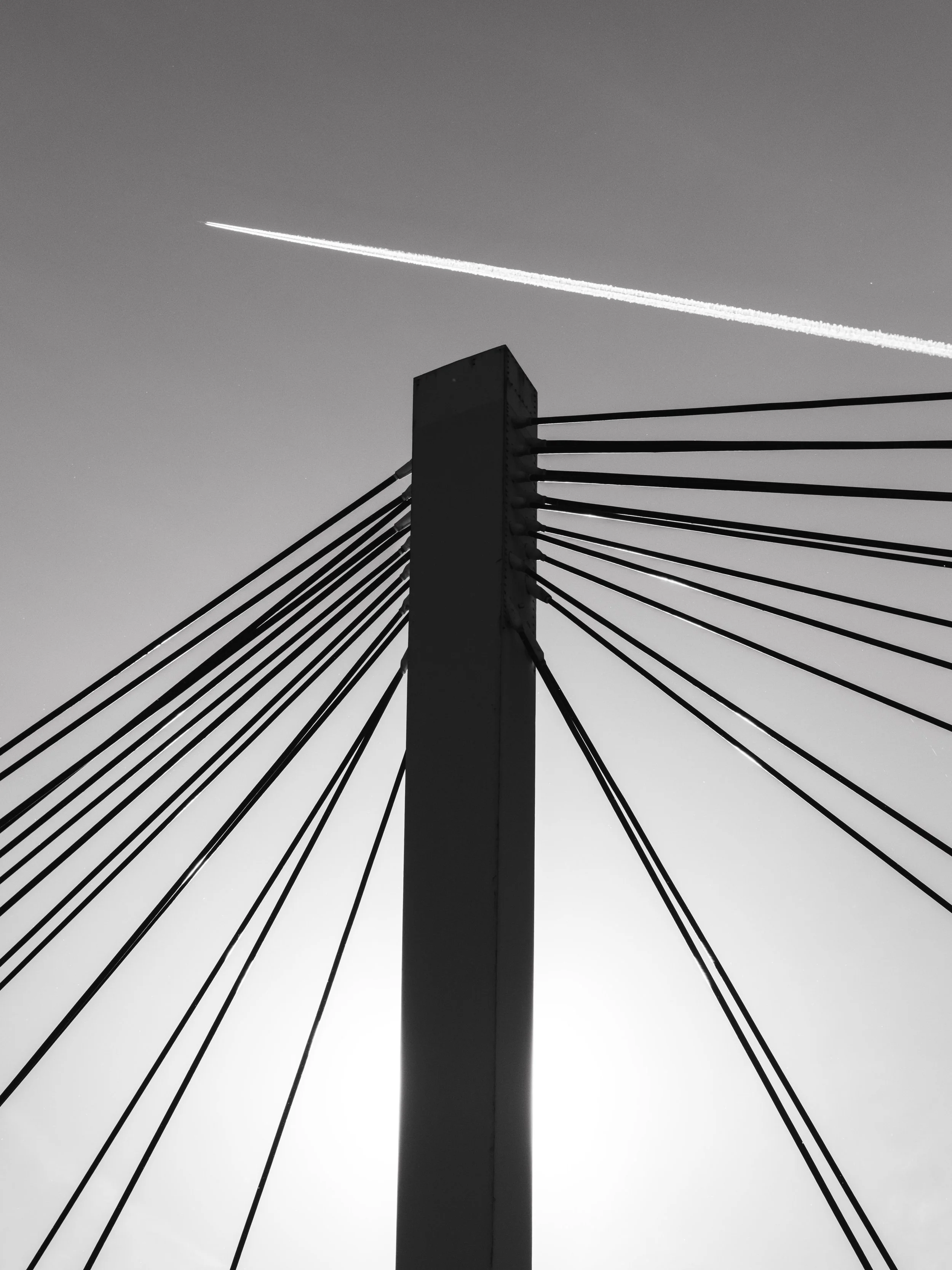 Schwarz-Weiß-Foto einer Brücke mit Kabeln und einer hohen Säule, im Himmel ein Flugzeug mit Kondensstreifen.