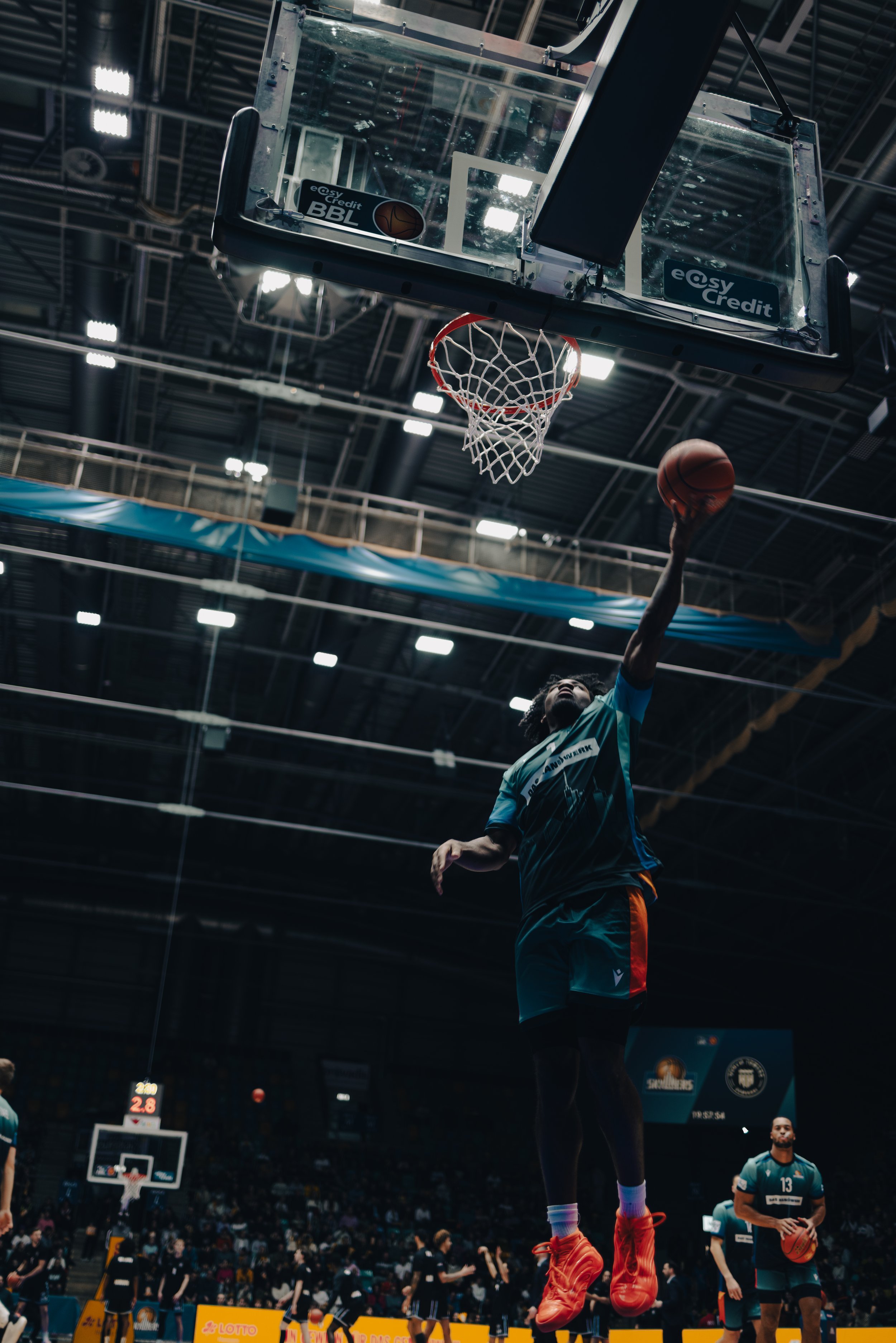Basketballspieler springt zum Korb, Basket im Hintergrund, Indoor-Basketballhalle, Publikum im Hintergrund, dunkle Beleuchtung, Sportbekleidung, Fokus auf Spieler in Aktion. Sportfotografie bei den Frankfurt Skyliners.