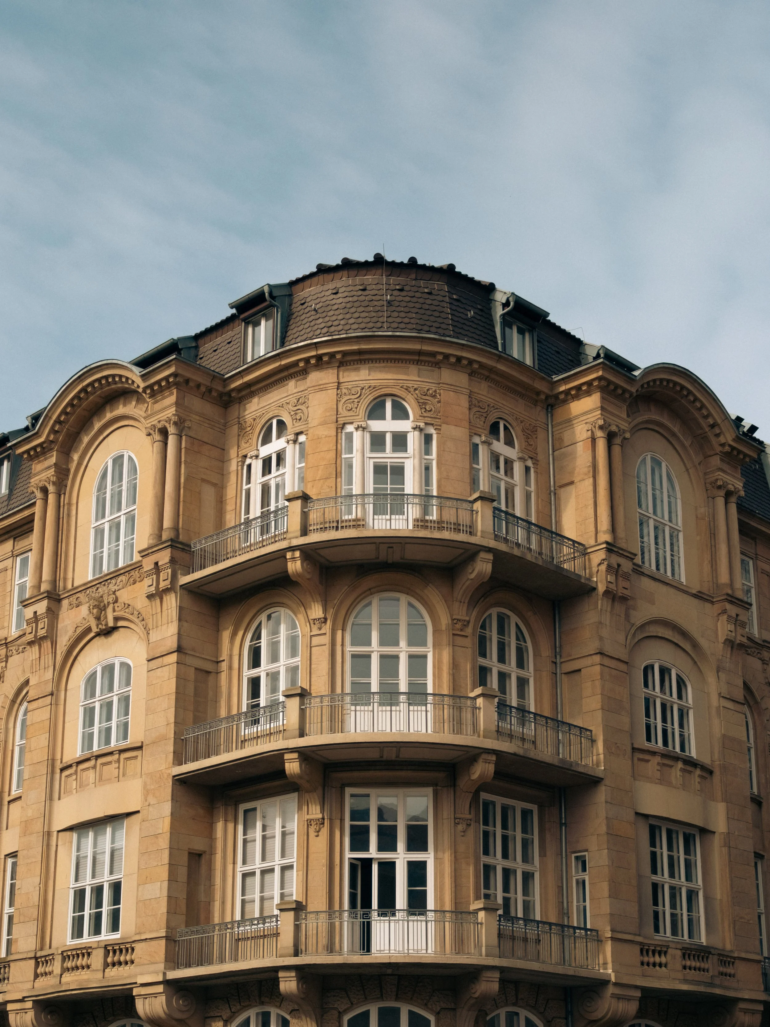 Auf der Aufnahme ist ein historisches, mehrstöckiges Gebäude mit einer auffälligen, in beige Tönen gehaltenen Fassade und großen Fenstern zu sehen. Es verfügt über mehrere Balkone mit dekorativen Geländern, und die Architektur ist im Stil des späten 