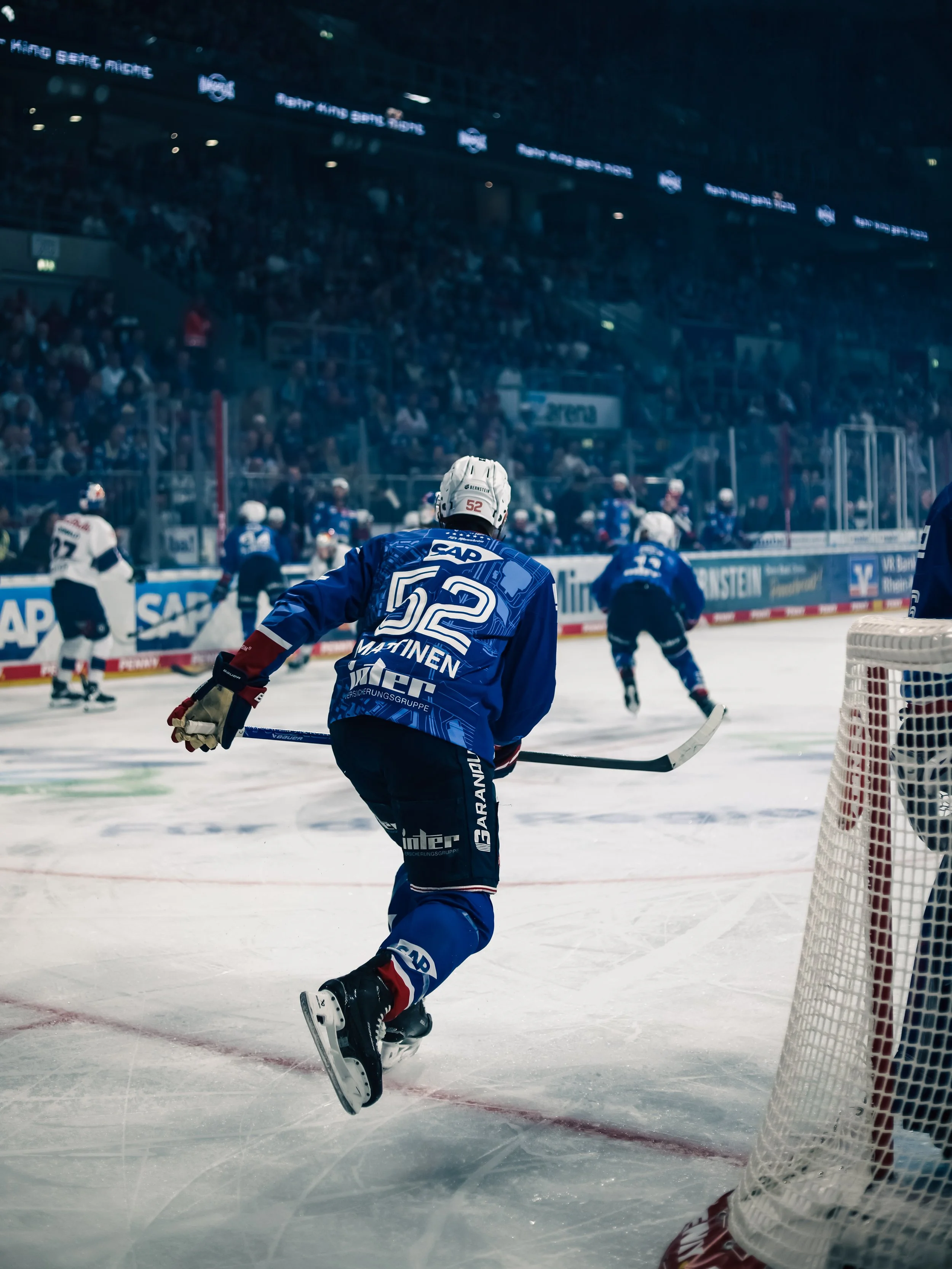 Eishockeyspieler der Adler Mannheim im blauen Trikot mit der Nummer 52 vor dem Tor, in einem Stadion mit einer vollen Zuschauertribüne im Hintergrund. Nick Mattinen. Sportfotografie bei den Adler Mannheim in der Deutschen Eishockey Liga.