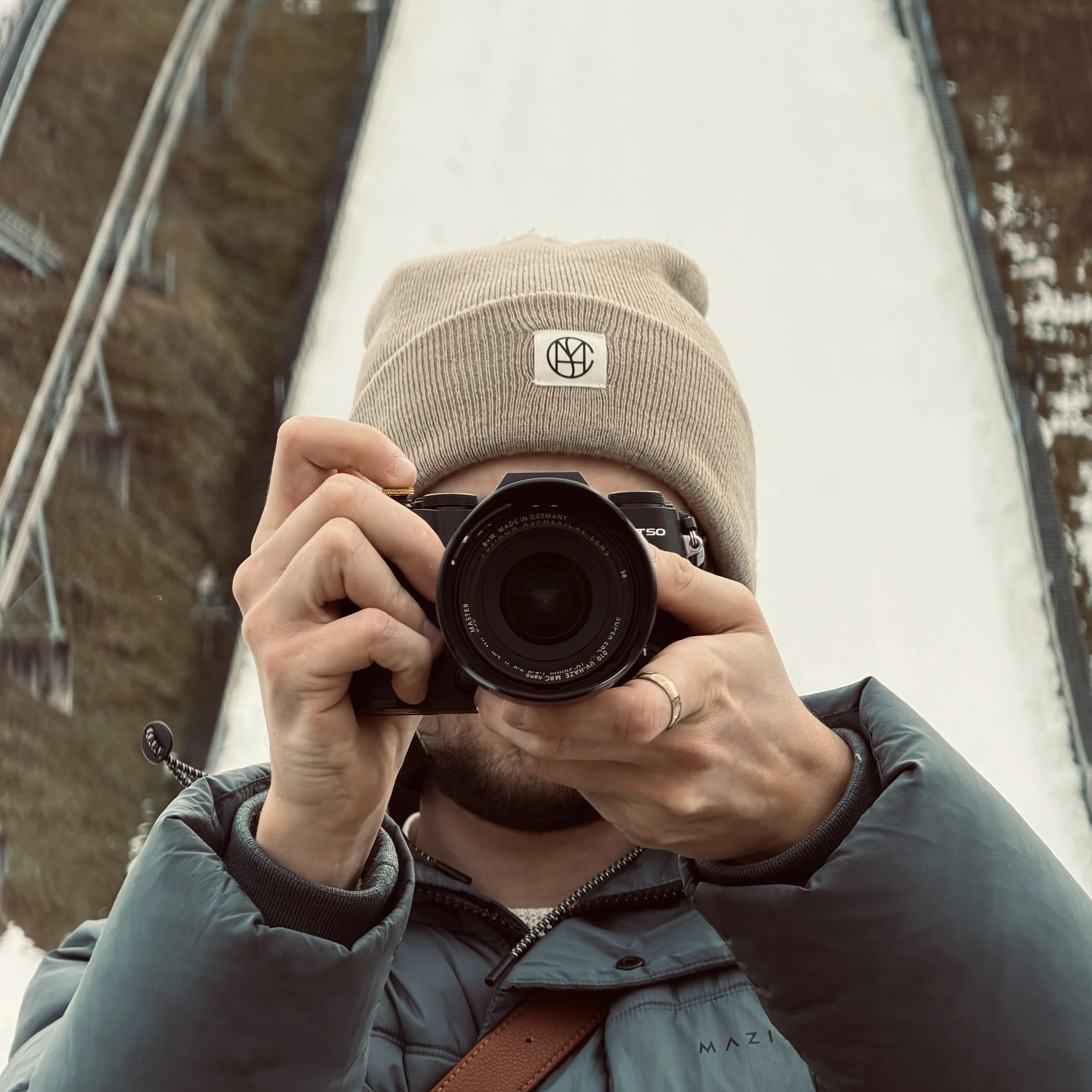 Fotograf der Seite trägt eine braune Mütze mit einem schwarzen Logo, hält eine Kamera und macht ein Selbstporträt vor der Skisprungschanze in Garmisch-Patenkirchen.