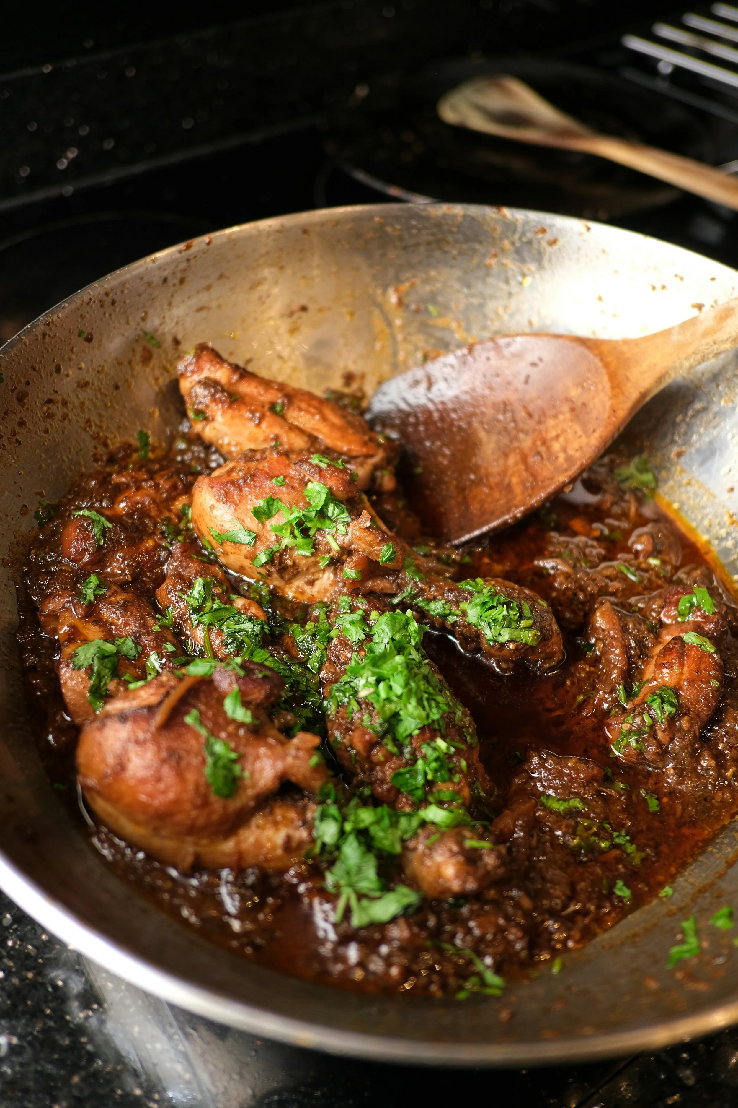 Cooked chicken pieces in a dark sauce with chopped fresh herbs on top, in a frying pan.