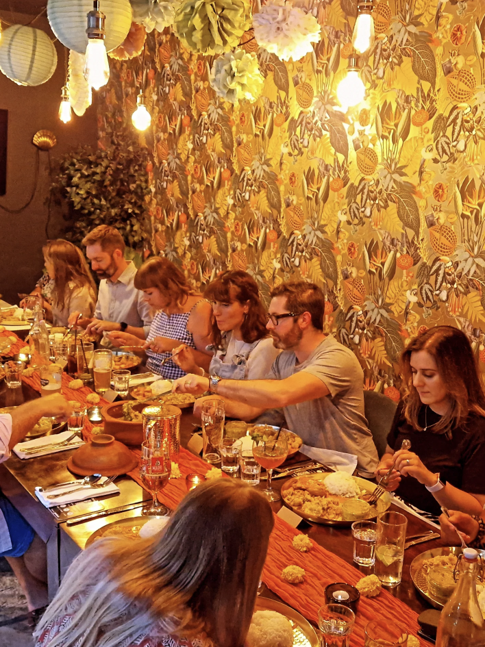 Group of people dining together at a long table in a restaurant with tropical leaf and fruit patterned wallpaper, hanging paper lanterns, and warm lighting.