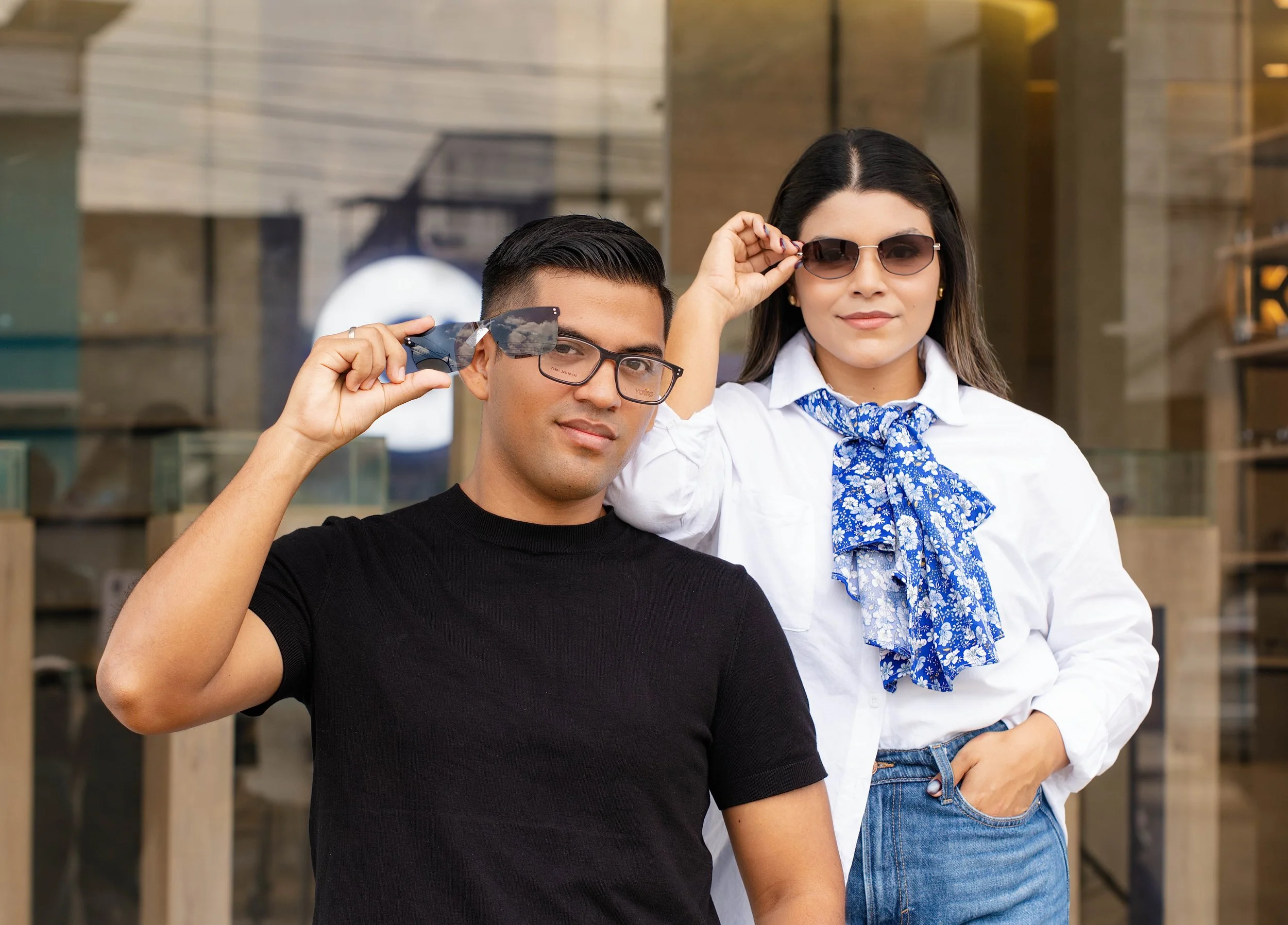 Jóvenes con gafas, el hombre con camiseta negra y la mujer con blusa blanca y pañuelo azul, posando en un lugar urbano.