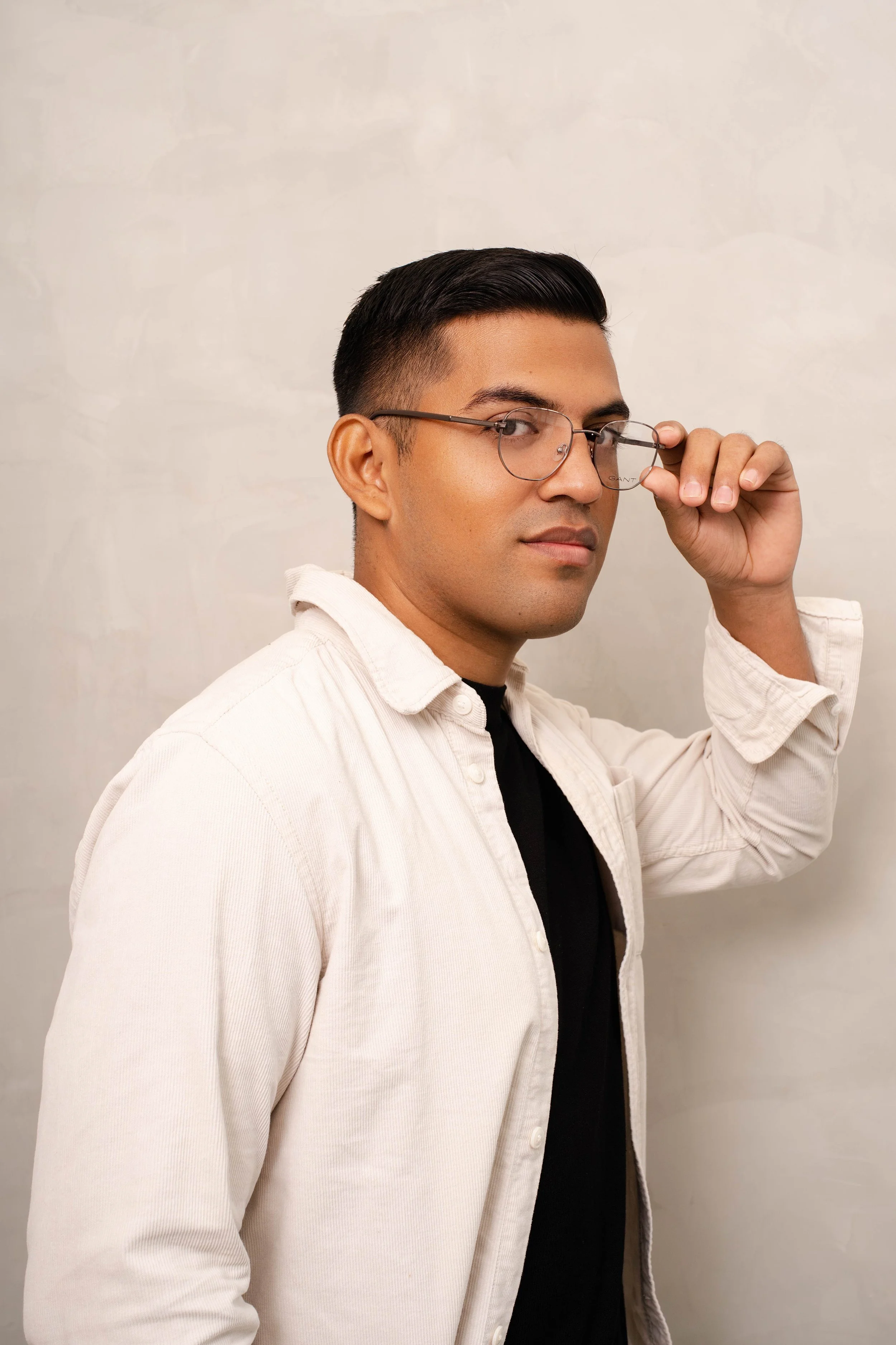 Hombre con gafas, cabello corto y oscuro levantando las gafas con la mano derecha en un fondo liso y claro.