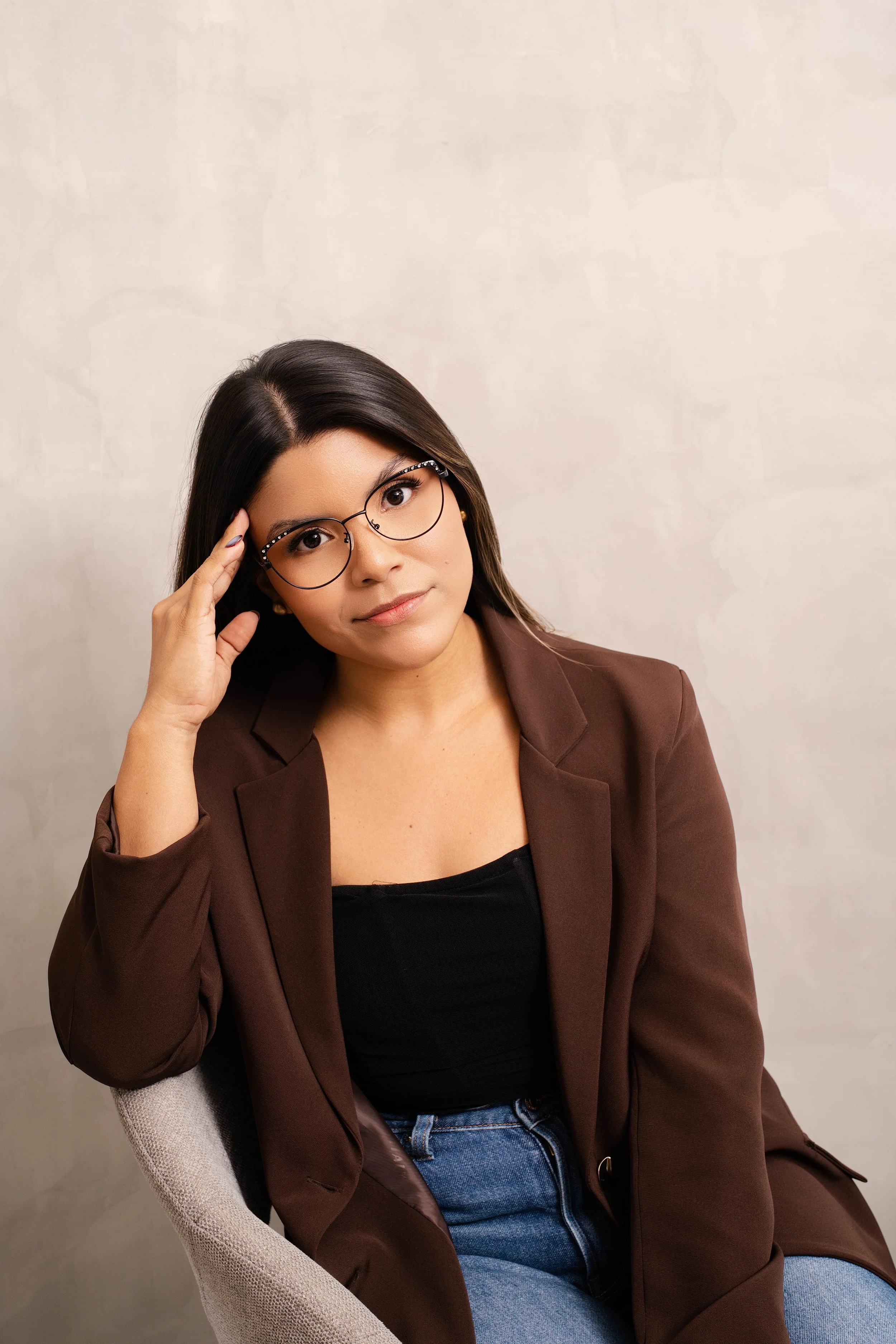 Mujer con gafas, cabello oscuro, usando blazer marrón y camiseta negra, sentada en una silla gris, con fondo neutro.