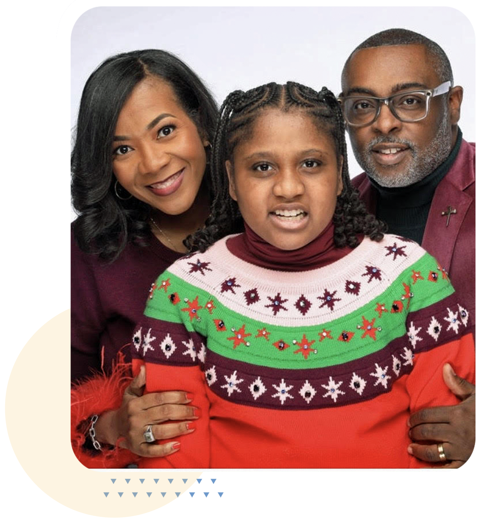 A family of three people, a woman, a girl, and a man, smiling and posing together against a plain white background. The girl is wearing a colorful Christmas sweater with red, green, white, and black patterns.