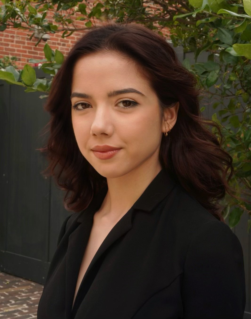 A woman with wavy, shoulder-length dark hair and light skin, dressed in a black blazer, standing outdoors in front of green foliage and a brick wall.