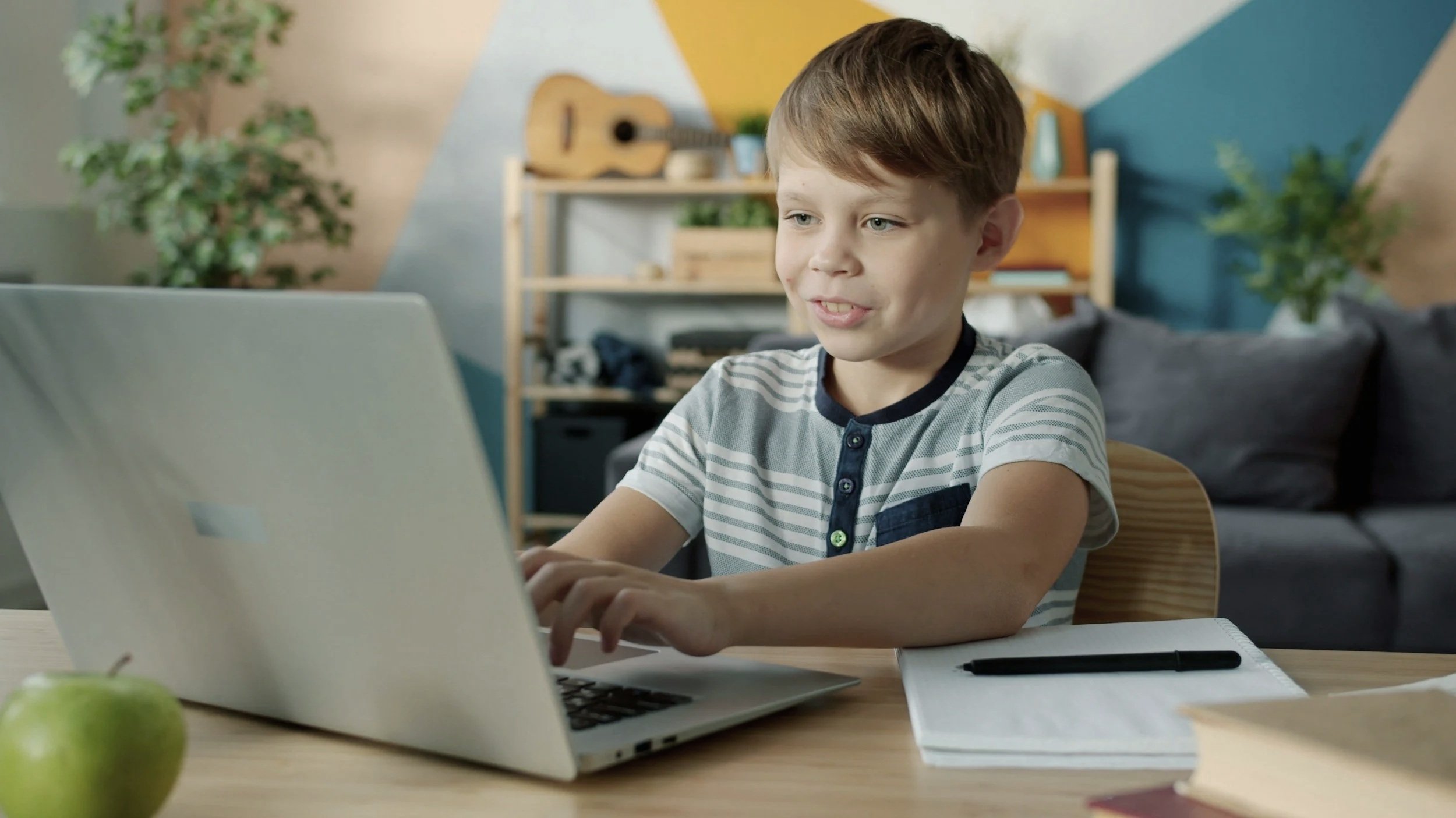 A young boy with light brown hair sitting at a wooden table, using a laptop, with a green apple, notebook, and pen in front of him in a room with colorful geometric decor, a bookshelf with a guitar, plants, and a gray sofa in the background.