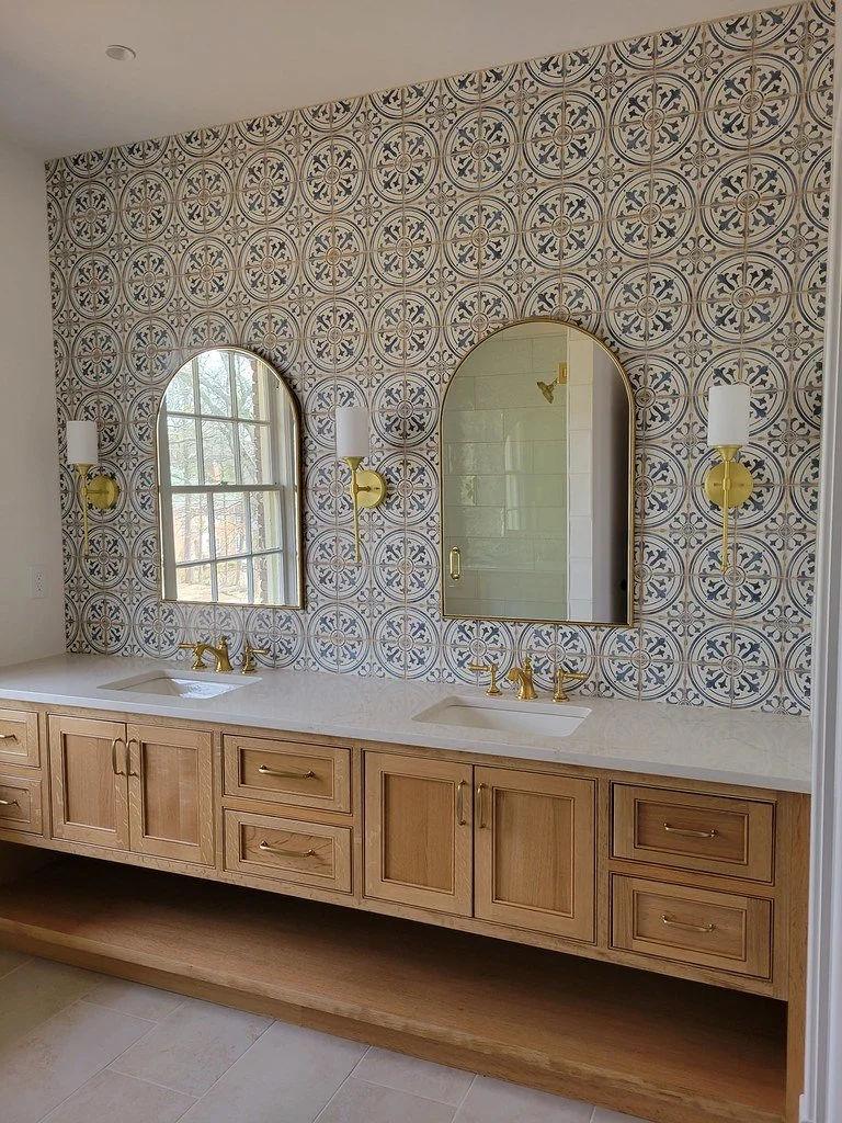 Bathroom vanity with wooden cabinets, two sinks, gold fixtures, a large mirror, and decorative patterned tile backsplash.