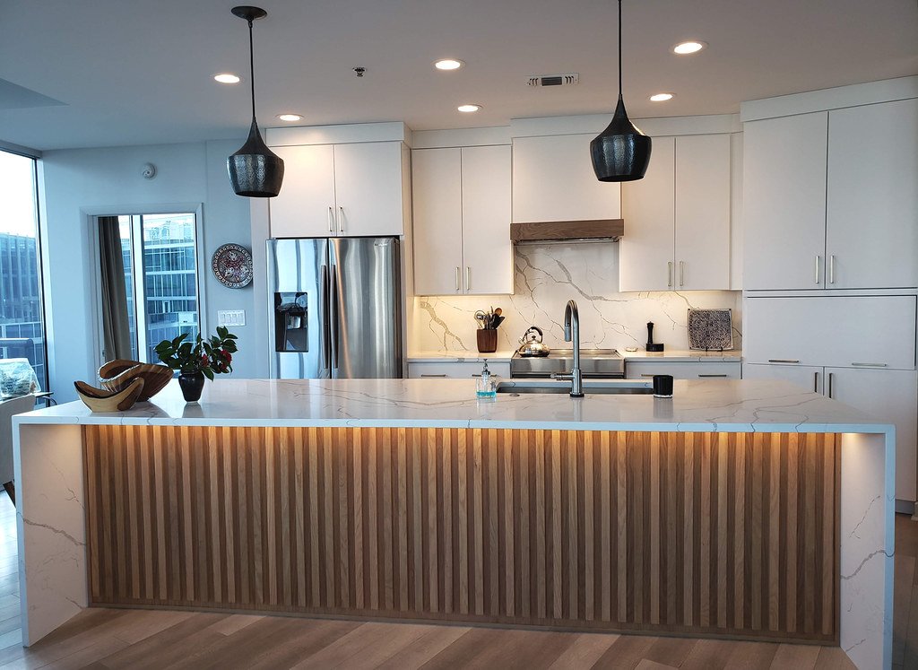 Modern kitchen with white cabinetry, marble backsplash, stainless steel refrigerator, and an island with wood paneling and built-in lighting. Pendant lights hang above the island.