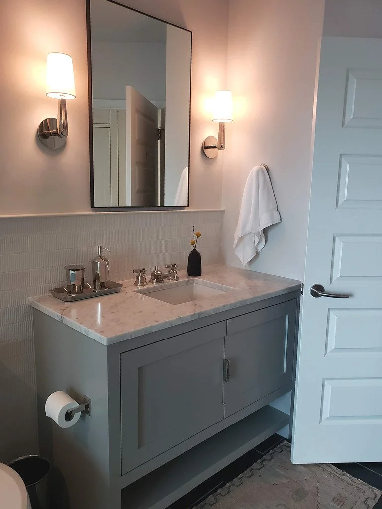 Bathroom vanity with a marble countertop, a small sink, a mirror, two wall-mounted lights, a towel hanging on a hook, and a doorway.