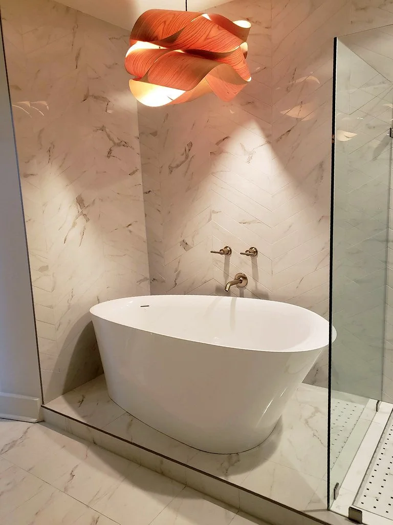 A modern bathroom with a freestanding oval bathtub on a marble platform, marble walls, a glass shower enclosure, and a contemporary wooden pendant light.