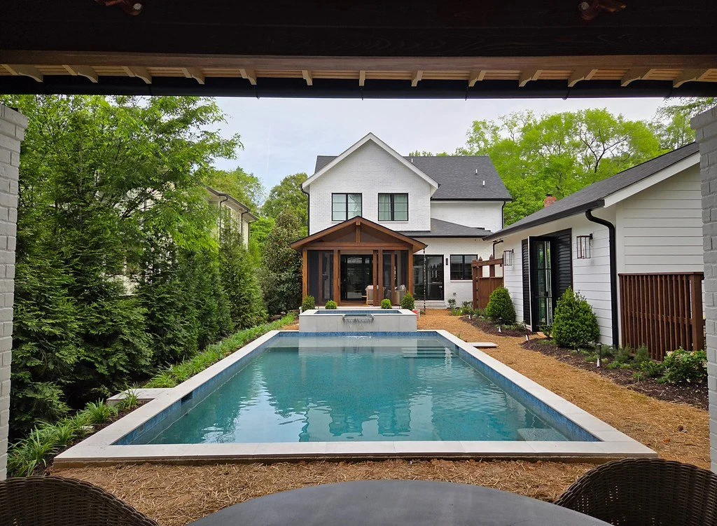 View of a backyard with a rectangular swimming pool, surrounded by a landscaped garden with bushes and trees, and a white house with a porch at the far end.