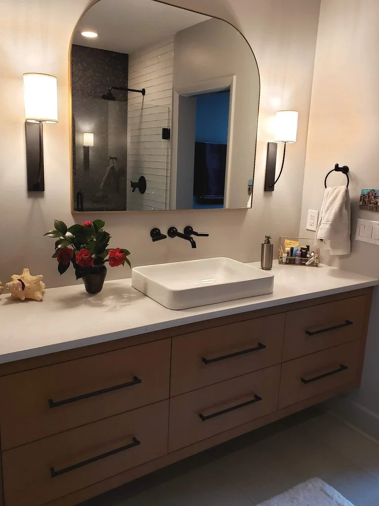 Modern bathroom vanity with a white vessel sink, black fixtures, a large mirror, two wall-mounted lights, a potted plant with red flowers, a shell, toiletries, and a towel.