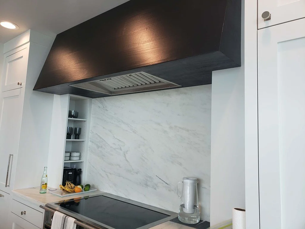 Kitchen with white cabinets, marble backsplash, and black range hood. Items include a water pitcher, glass bottle, and fruit on the counter.