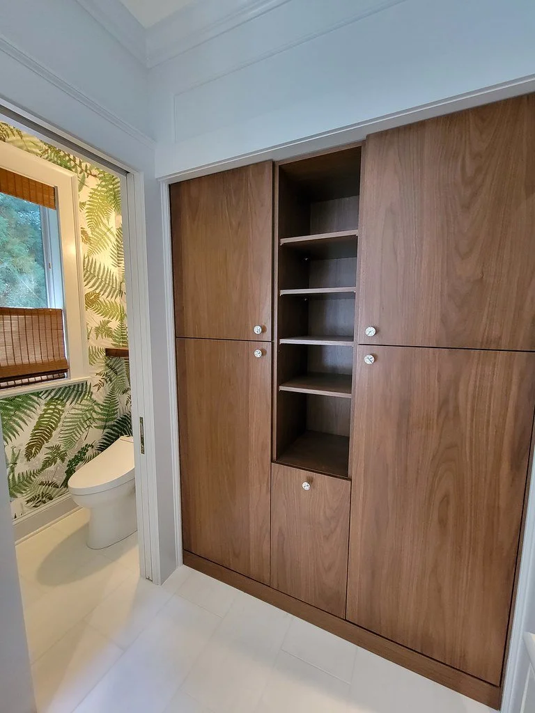 A wooden built-in cabinet with open shelves in a corner of a room, next to a doorway leading to a bathroom with a fern-patterned wallpaper and a toilet.
