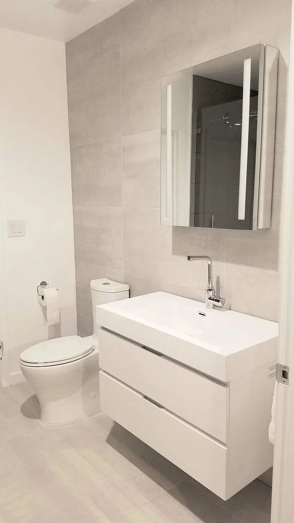 Modern bathroom with white toilet, white vanity sink with a sleek faucet, gray tiled wall, and a mirrored medicine cabinet.