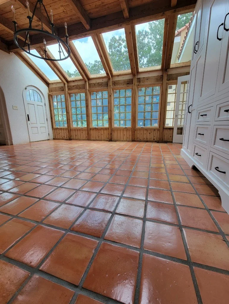 A bright sunroom with large glass windows, a wooden ceiling with exposed beams, and terracotta tile flooring. There is a white cabinet on the right and an overhead chandelier on the left.