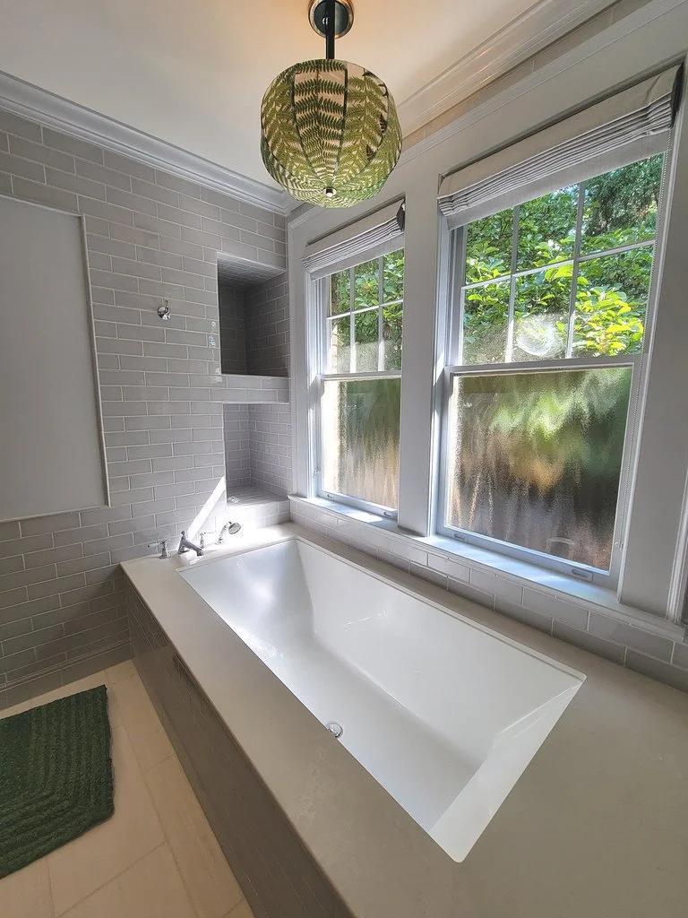 A white bathtub beside two large frosted and clear windows in a bathroom, with a green woven lampshade hanging from the ceiling and a green textured bath mat on the floor.