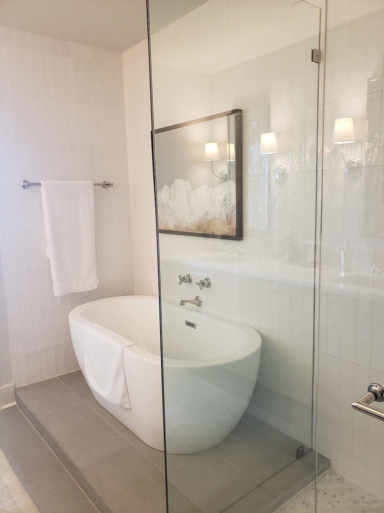 Modern bathroom with a white bathtub behind a glass partition, a towel hanging on a rail, and a framed mountain landscape picture on the wall with wall-mounted lamps reflected in the glass.