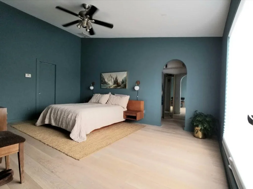 Bedroom with blue walls, beige bed with pillows, wall art, two white wall sconces, wooden nightstand, ceiling fan, potted plant, and light wood flooring.