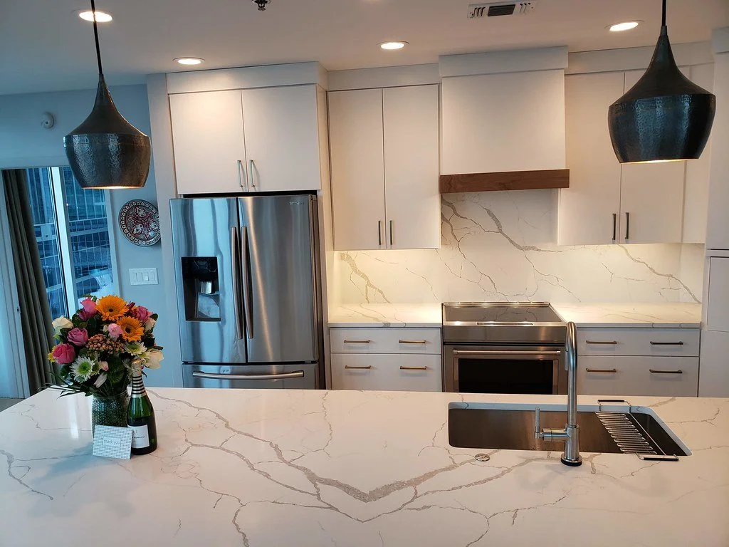 Modern kitchen with white marble countertops and backsplash, stainless steel refrigerator and oven, black pendant lights, a bouquet of colorful flowers, and a small card and bottle on the island.