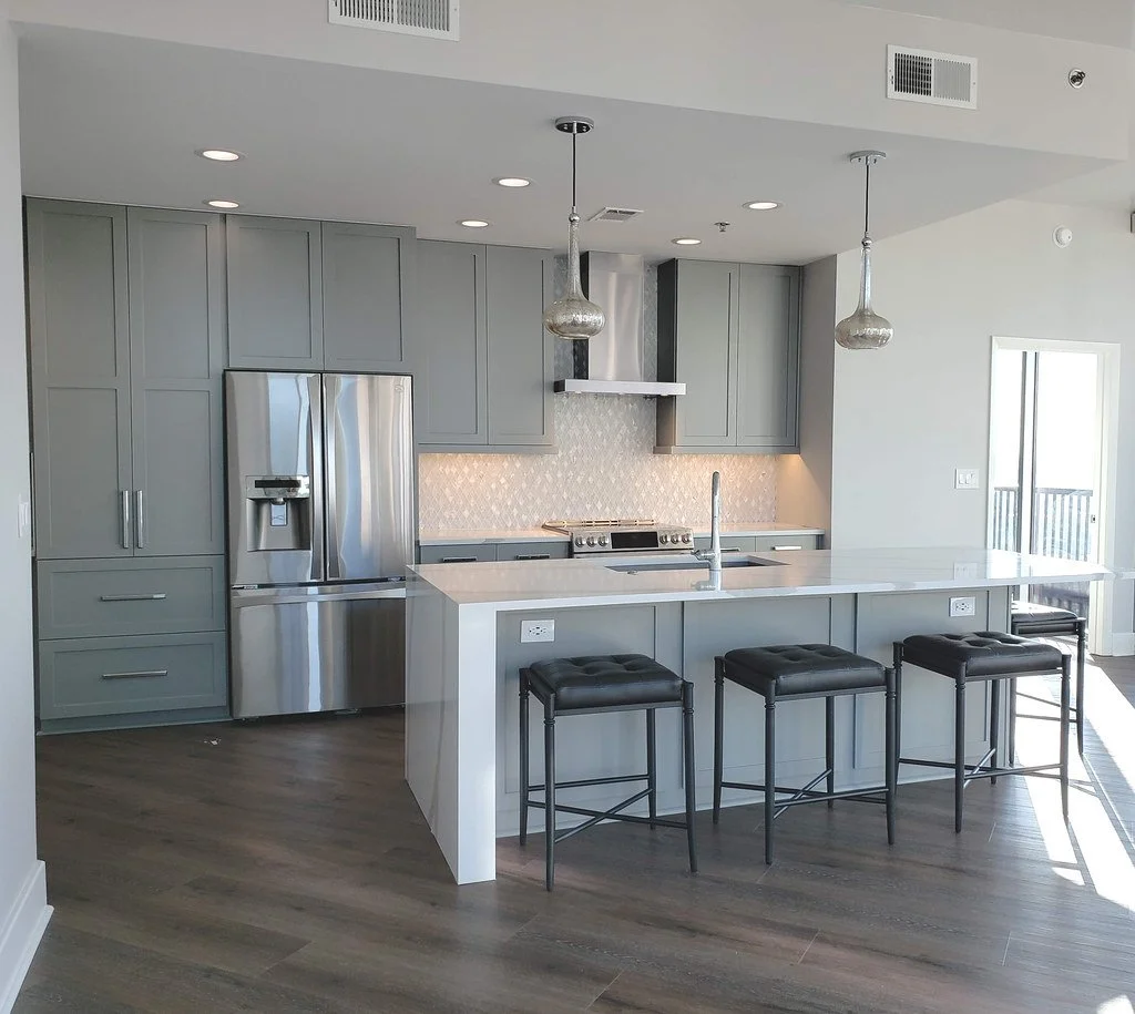 Modern kitchen with gray cabinets, stainless steel refrigerator, combined oven and cooktop, white marble countertop island with three black barstools, pendant lights, and hardwood flooring.