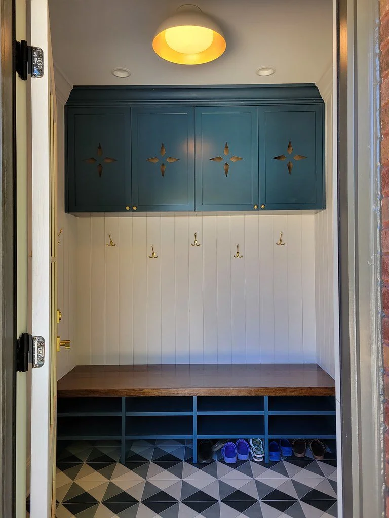 Entryway mudroom with blue cabinets, gold hooks, a wooden bench, cubby storage, and shoes on the floor with geometric black and white tile.
