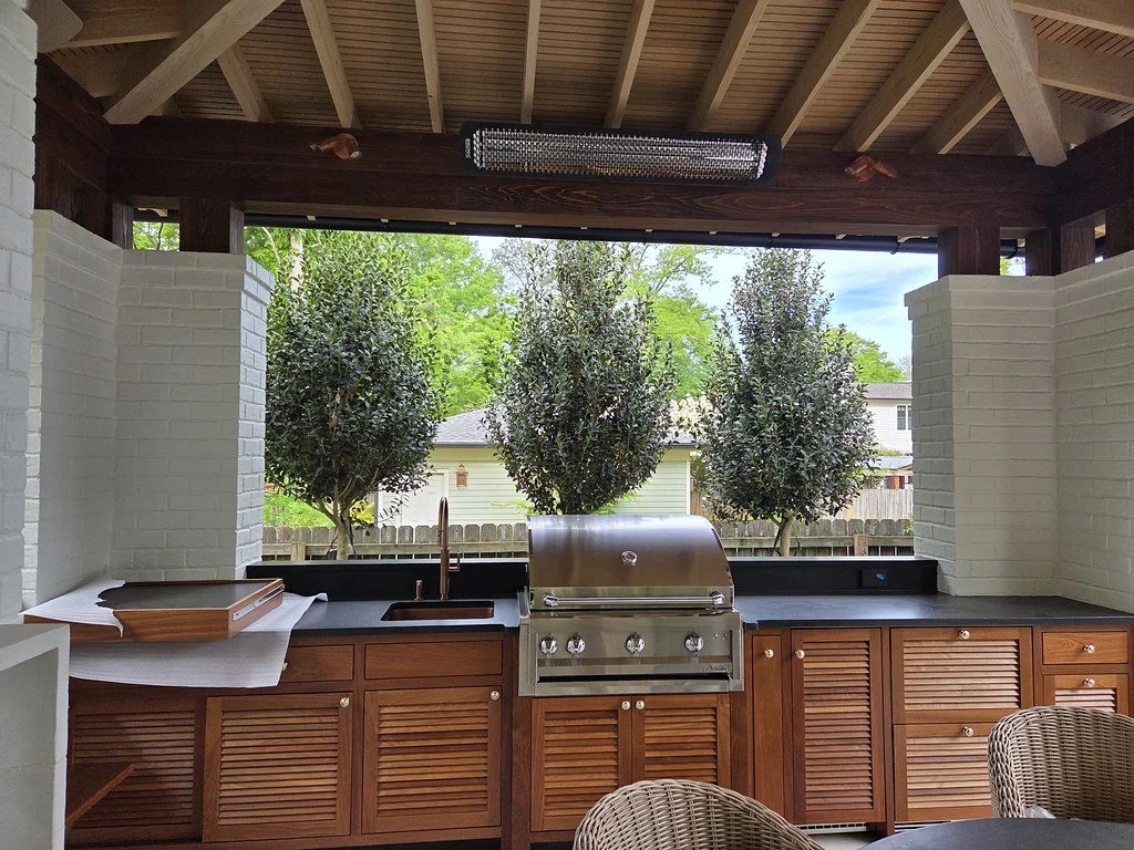 Outdoor kitchen with a built-in grill, wooden cabinets, a black countertop, a sink, and a view of green trees and a house beyond a white brick wall.