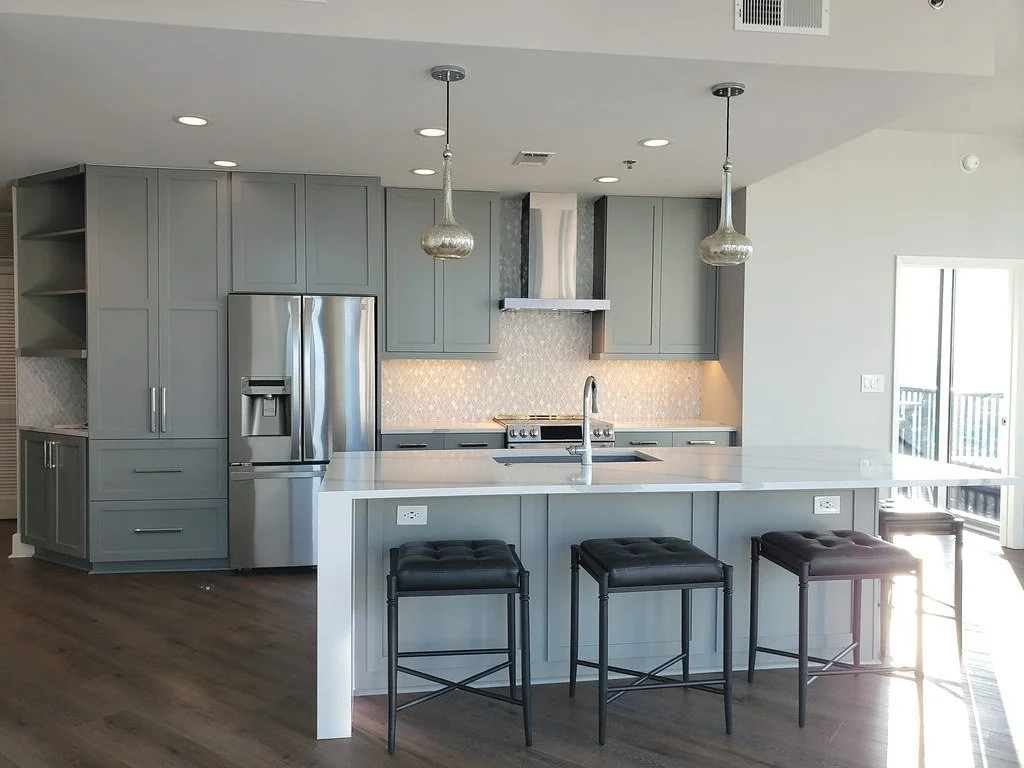 Modern kitchen with gray cabinets, stainless steel refrigerator, island with white countertop, black barstools, pendant lights, and a sliding glass door leading to an balcony.