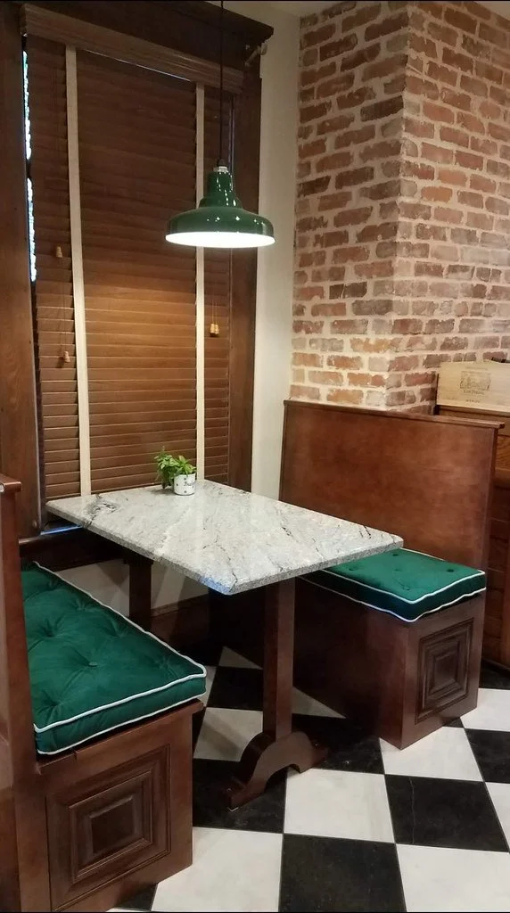 A cozy breakfast nook featuring a marble-topped table with two wooden booth seats with green cushions. A green hanging light fixture provides illumination, with exposed brick wall creating a warm ambiance.