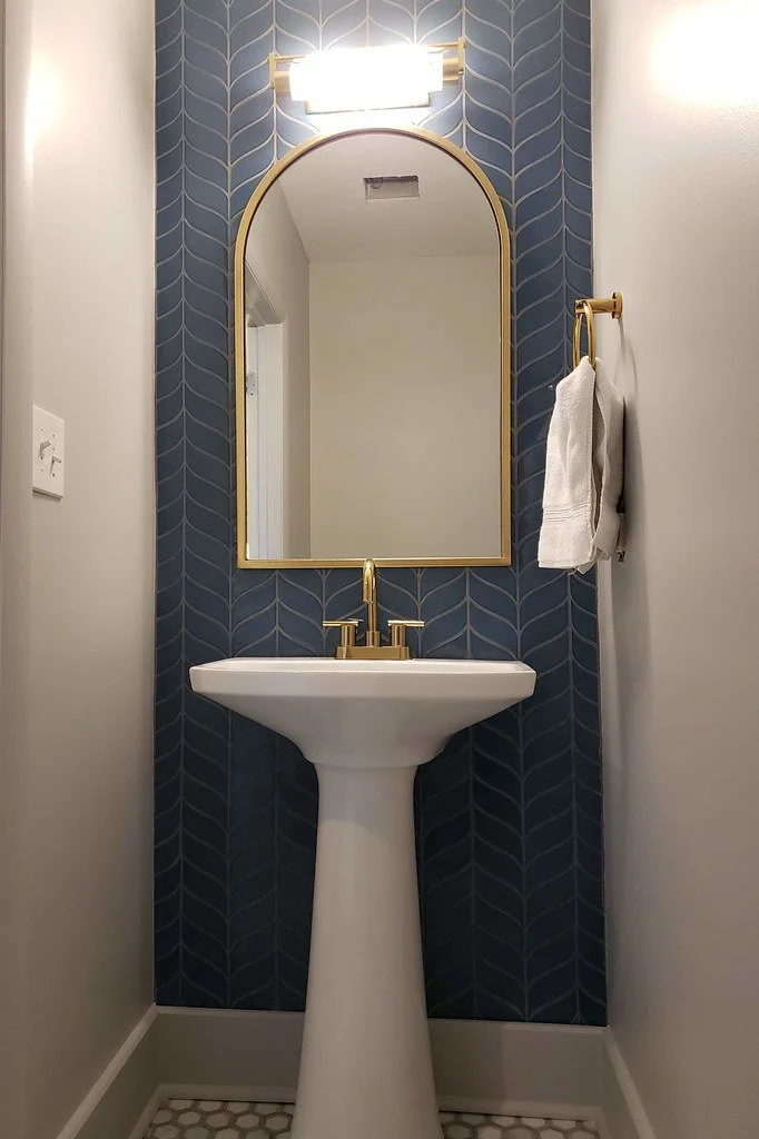 A small bathroom corner with a pedestal sink, gold faucet, white towel on a gold towel ring, large mirror with gold trim, and wall-mounted light fixture.