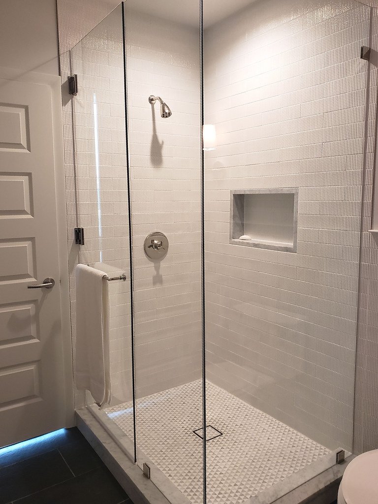 White tiled walk-in shower with glass enclosure, built-in niche, wall-mounted showerhead, and a towel bar outside the shower.