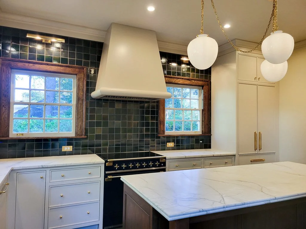 Kitchen with black tiled walls, white cabinets, a white marble countertop island, two windows with wooden frames, a white range hood, and three hanging globe pendant lights.