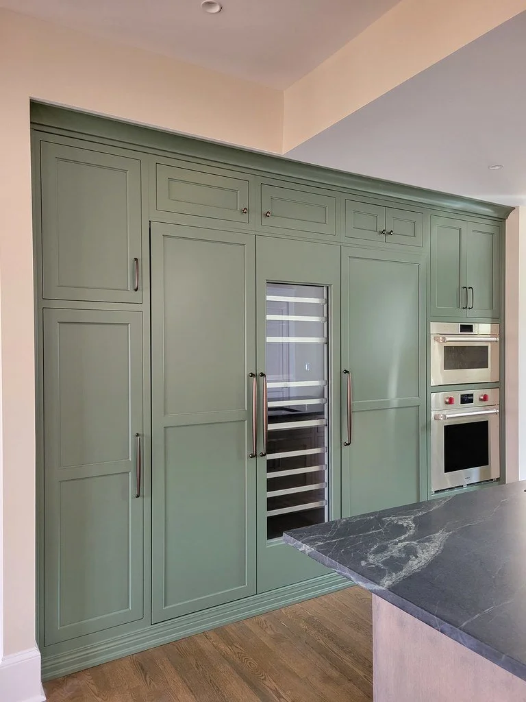 Green kitchen cabinets with built-in oven and a glass-front wine fridge, next to a dark marble countertop.