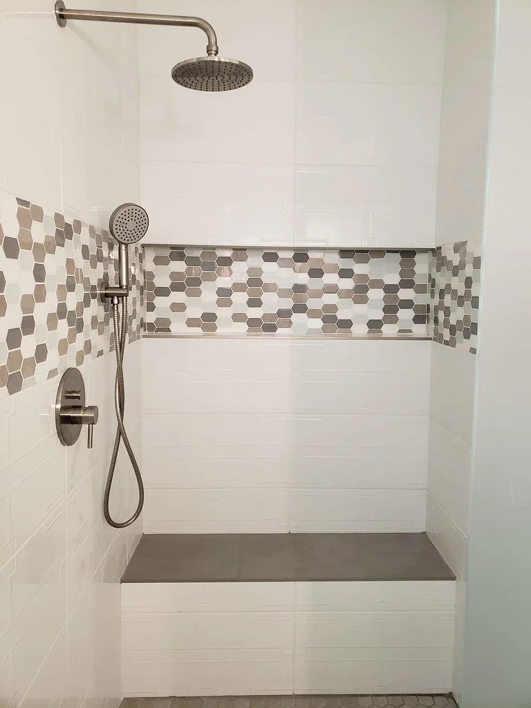 A modern walk-in shower with a large rainfall showerhead, a handheld shower, and gray and beige hexagonal tile accents on white tiled walls.