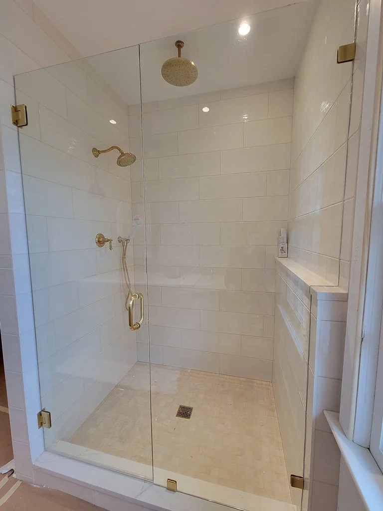 Glass-enclosed walk-in shower with beige tiles, a brass showerhead, handheld shower, and a built-in shelf on the right wall, with a drain in the center of the tile floor.