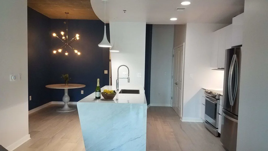 Modern kitchen with white island, stainless steel refrigerator, stove, and navy blue accent wall, with a dining area featuring a round table and a unique chandelier.