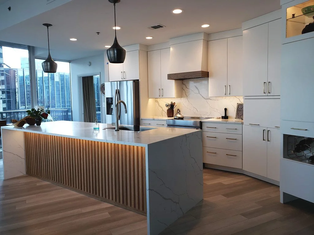 Modern kitchen with white cabinets, marble countertops, and a marble backsplash, featuring a large island with wooden slats and hanging black pendant lights.