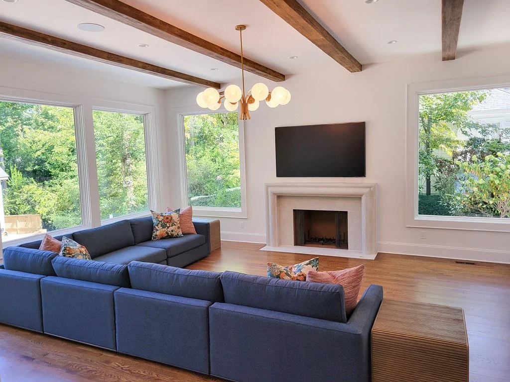 Living room with large windows, a gray sectional sofa with multiple pillows, a white fireplace with a mounted flat-screen TV above, wooden ceiling beams, a modern chandelier, and hardwood flooring.