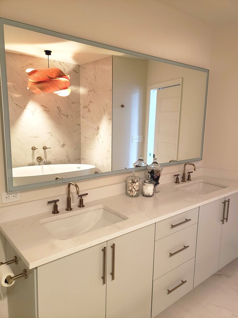 A double sink bathroom vanity with a large mirror, decorated with jars of shells and toiletries, and a marble-tiled bathtub with an overhead light fixture visible in the reflection.
