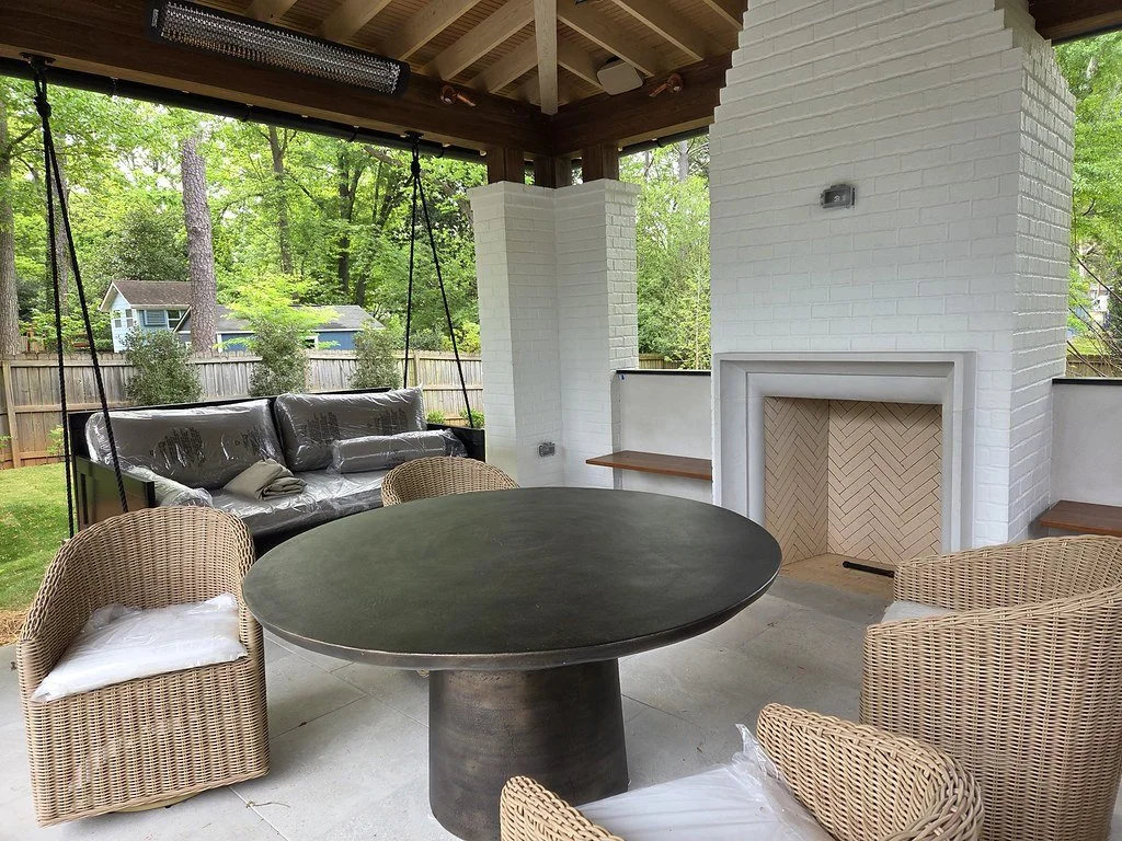 Covered outdoor patio with a white brick fireplace, wicker chairs with white cushions, a round black table, and a black sofa covered in plastic, surrounded by green trees and a backyard fence.