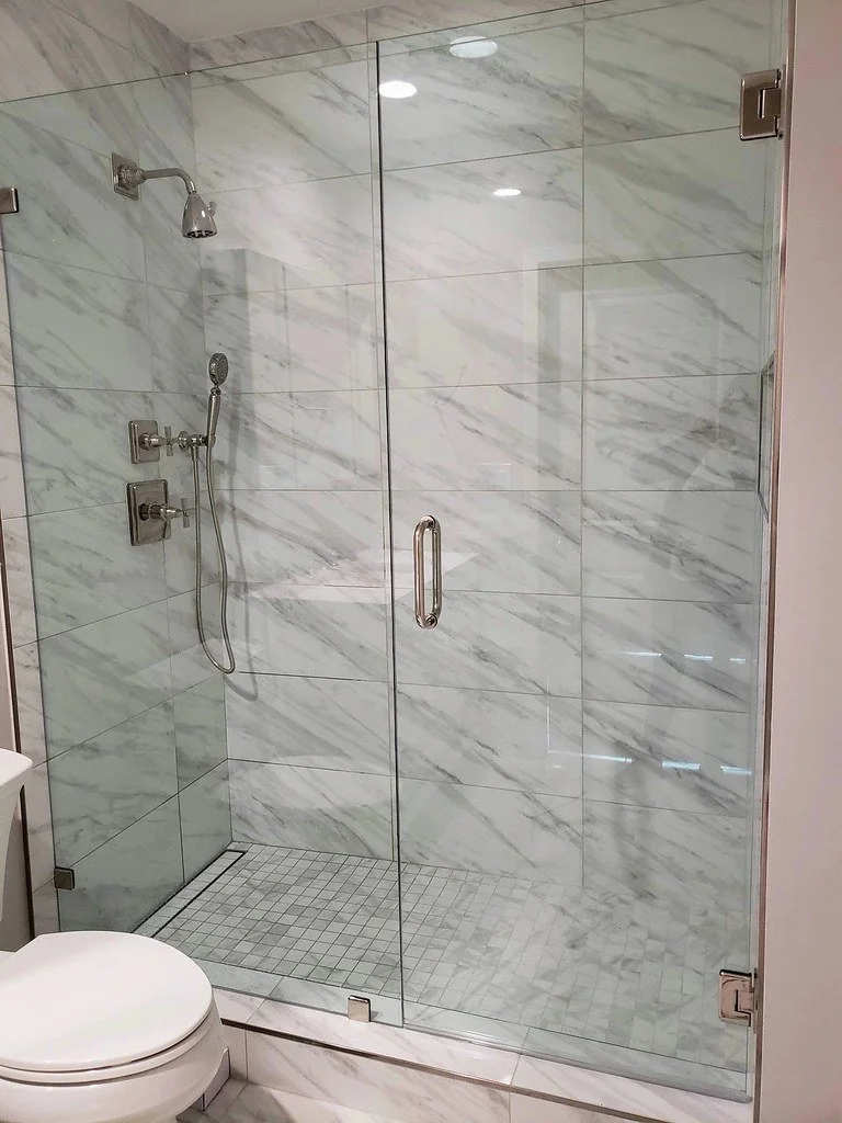 Modern walk-in shower with glass door and marble tiles, including a handheld showerhead and rain shower, in a bathroom with a toilet nearby.