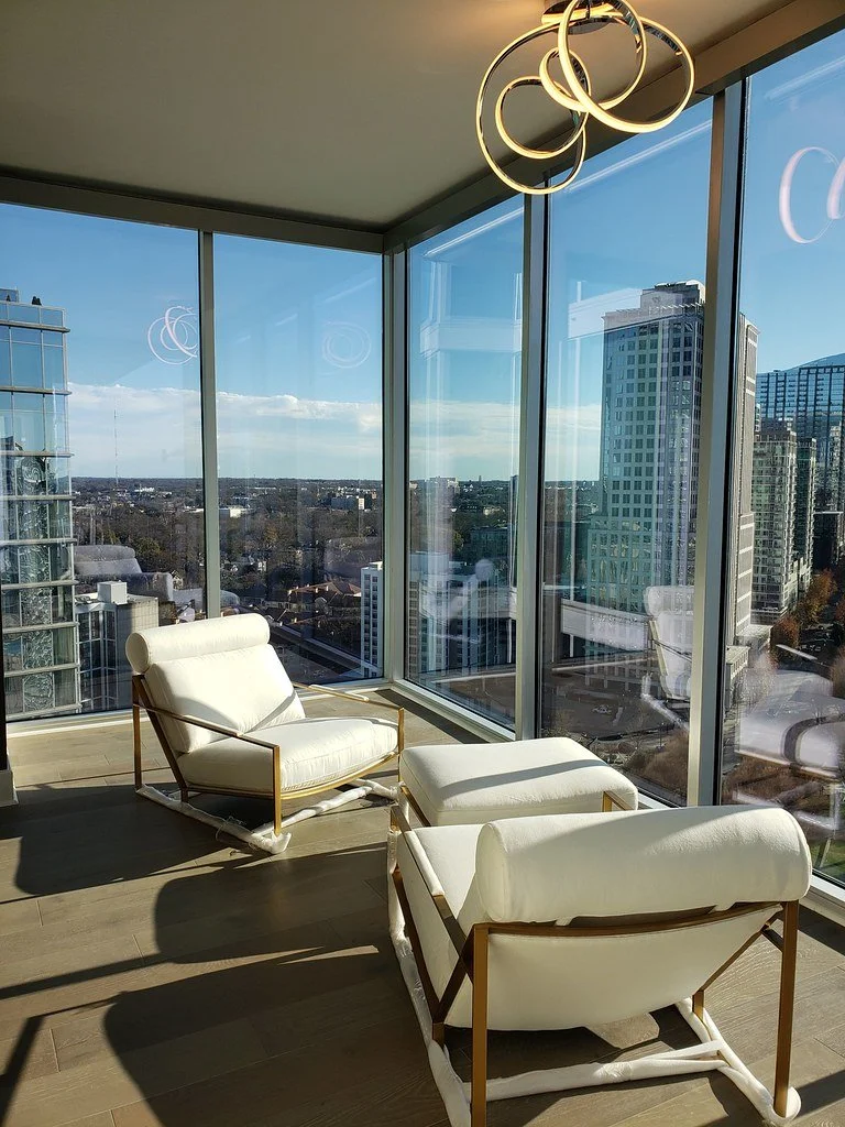 Modern high-rise apartment living room with floor-to-ceiling windows, two white lounge chairs with matching ottomans, and a unique circular ceiling light fixture, overlooking a cityscape with tall buildings and blue sky.