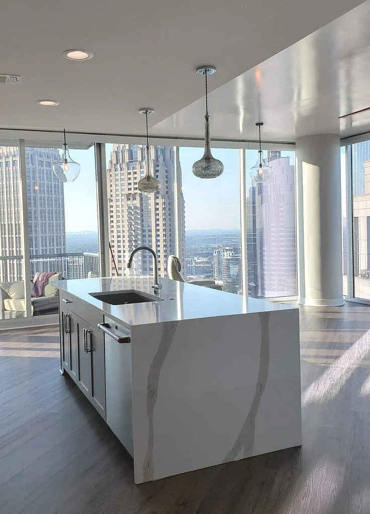 Modern kitchen island in a high-rise apartment with city skyline view, pendant lights, and large windows.